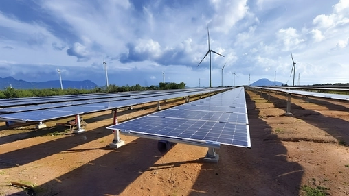 A solar farm with rows of photovoltaic panels and wind turbines in the distance under a partly cloudy blue sky