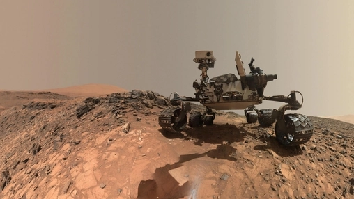 Six-wheeled Mars exploration rover on flat rocky Martian ground with a sand dune and hazy sky in the background